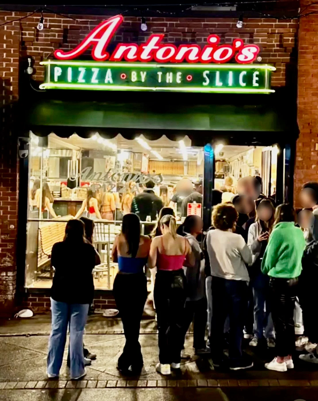Evening line outside Antonio's Pizza Amherst under the neon Pizza by the Slice sign