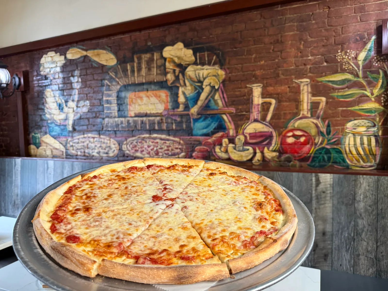 A whole cheese pizza on a tray in front of Antonio's pizzaiolo mural