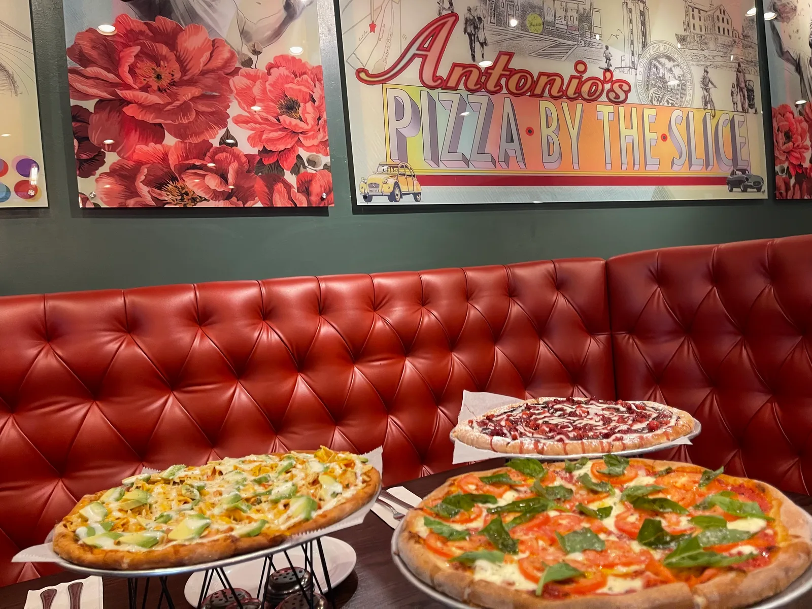 Three Antonio's pizzas on a red leather booth table beneath vintage brand posters